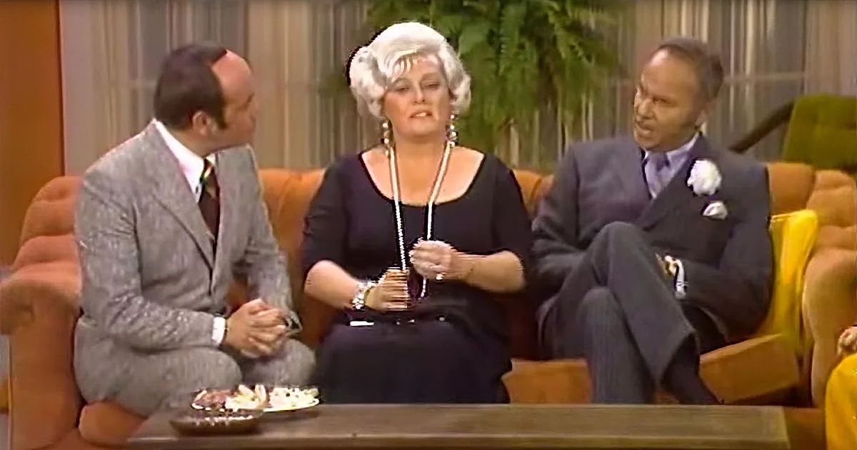 A woman in a black dress and white wig sits on a couch, holding a necklace, with two men in suits beside her.