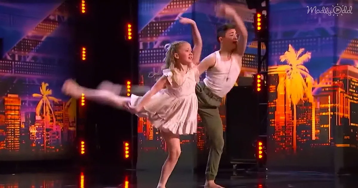 A young girl in a pink dress is dancing with a man on stage, both performing a ballet move against a cityscape backdrop.