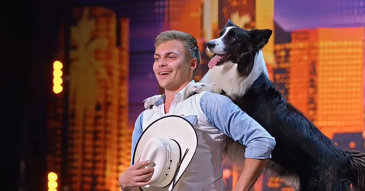 A man in a light blue shirt holds a white hat and has a black and white dog on his shoulders, smiling.
