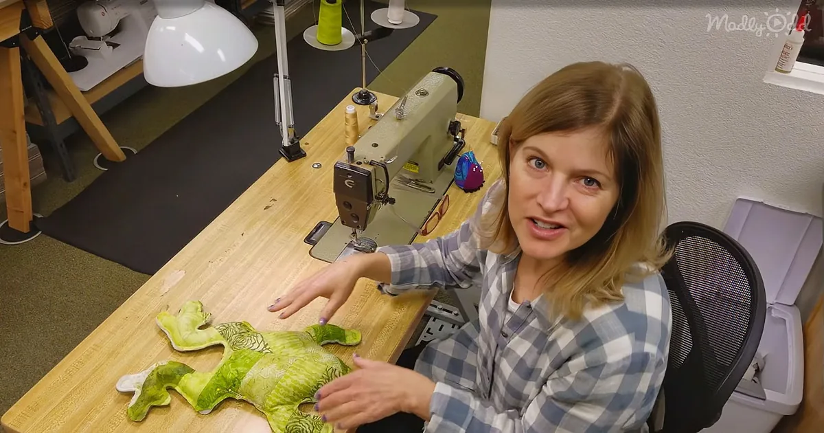 A woman sits at a sewing table with a green stuffed frog, surrounded by sewing equipment and supplies.