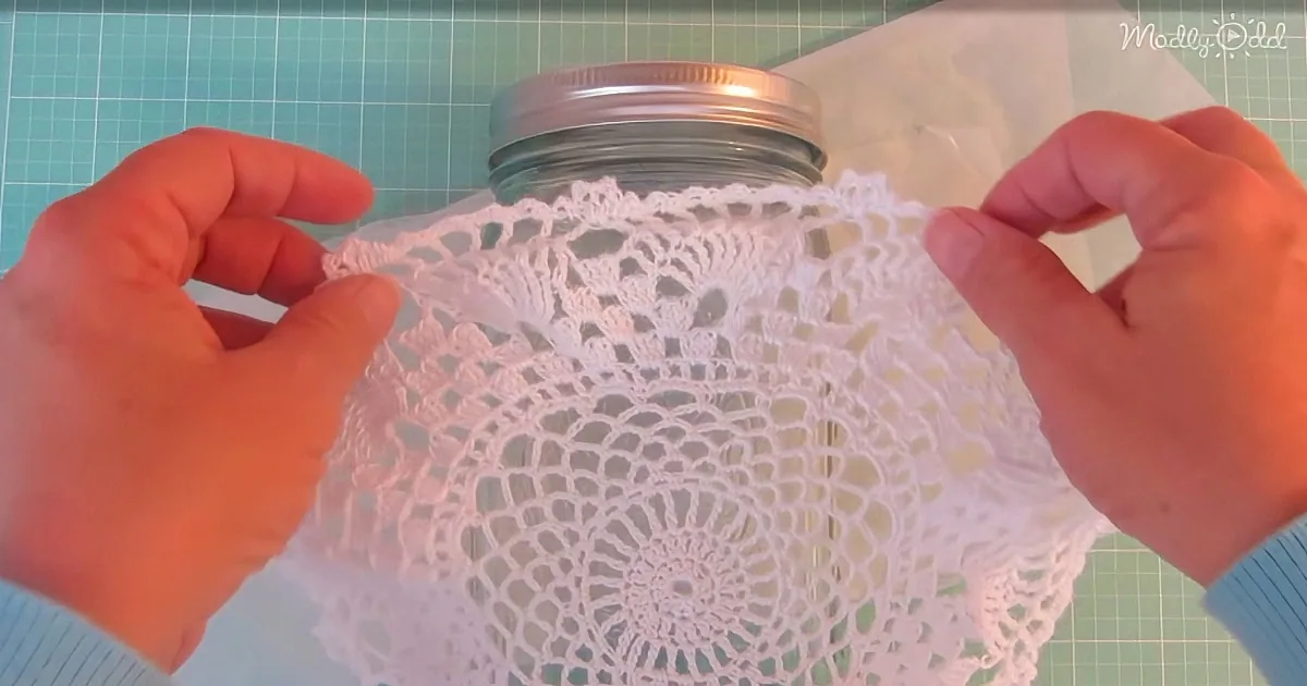 Hands hold a white doily over a glass jar on a green cutting mat.