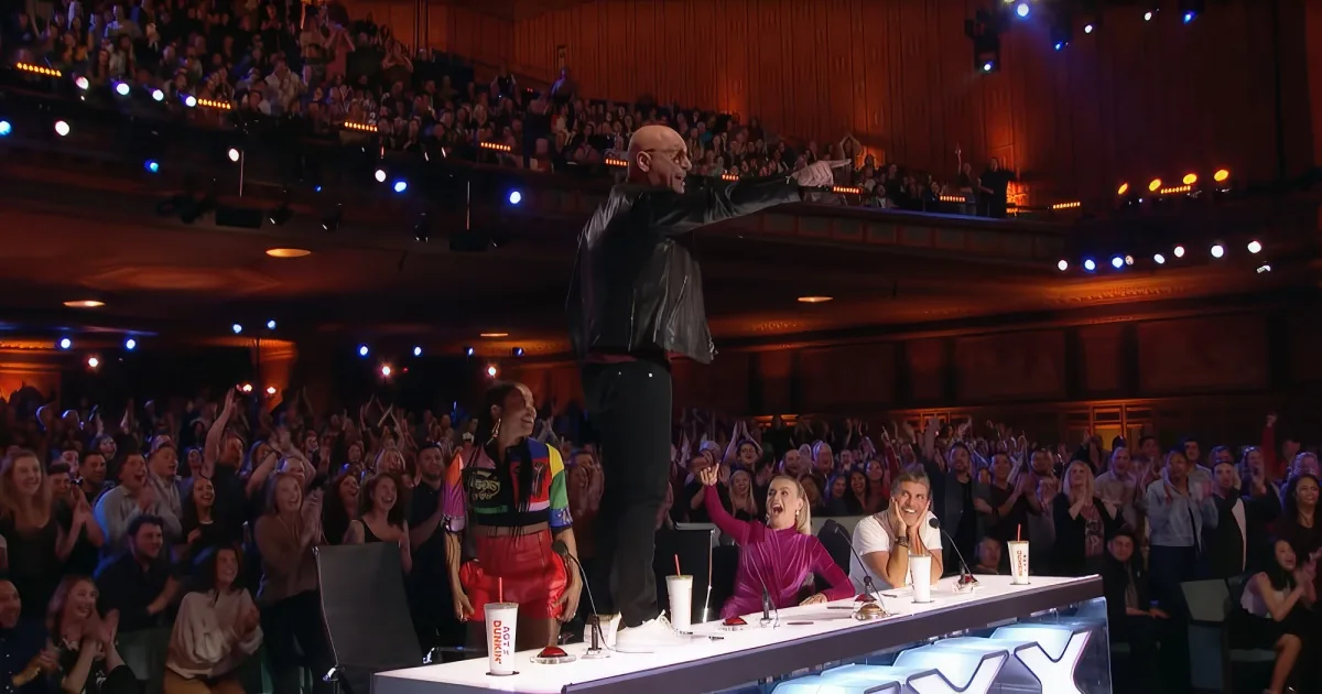 A man in a black leather jacket stands on stage, raising his arm, surrounded by an enthusiastic audience in a theater...