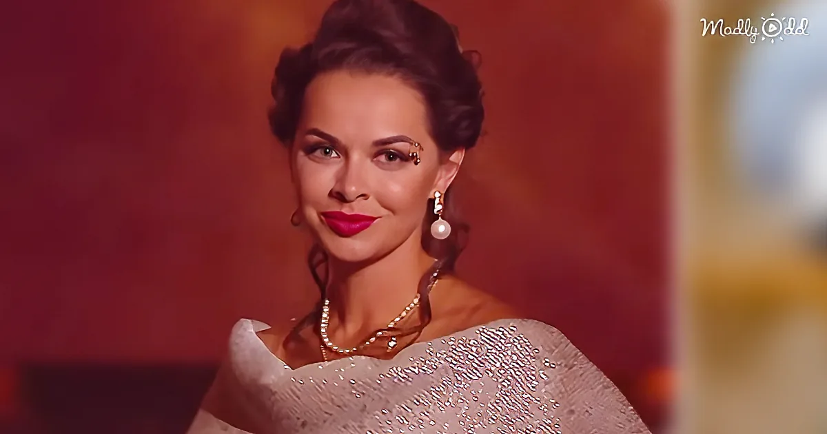 A woman in a sparkly dress with a pearl necklace and earrings, smiling with red lipstick.