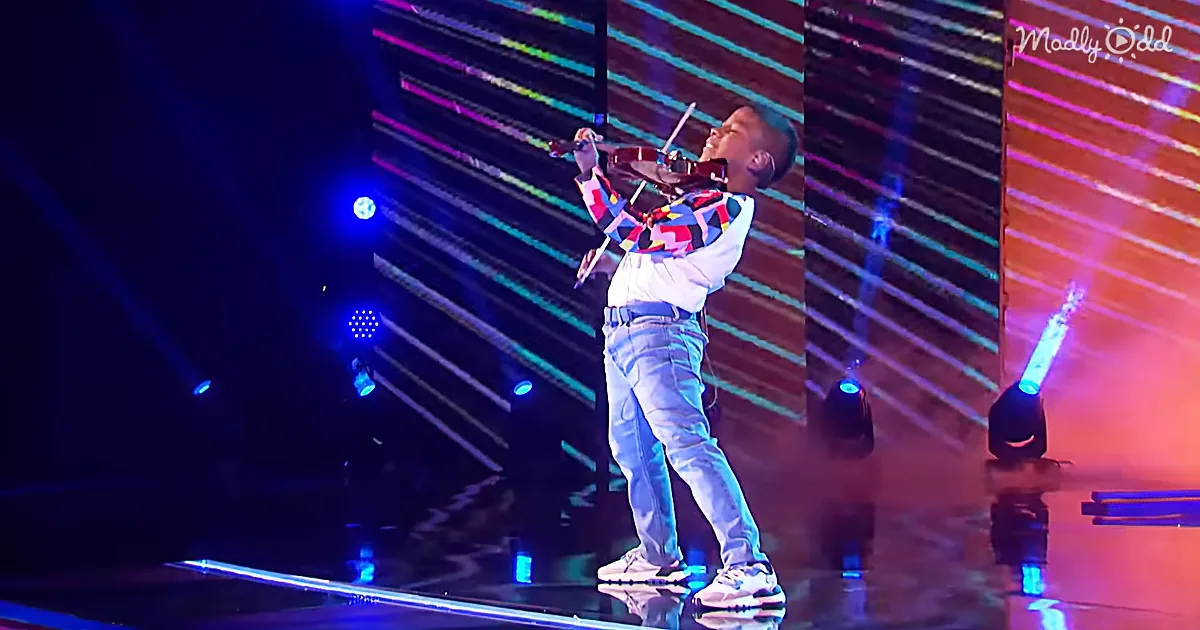 A young boy stands on stage, holding a violin, with colorful stage lights and a reflective floor behind him.