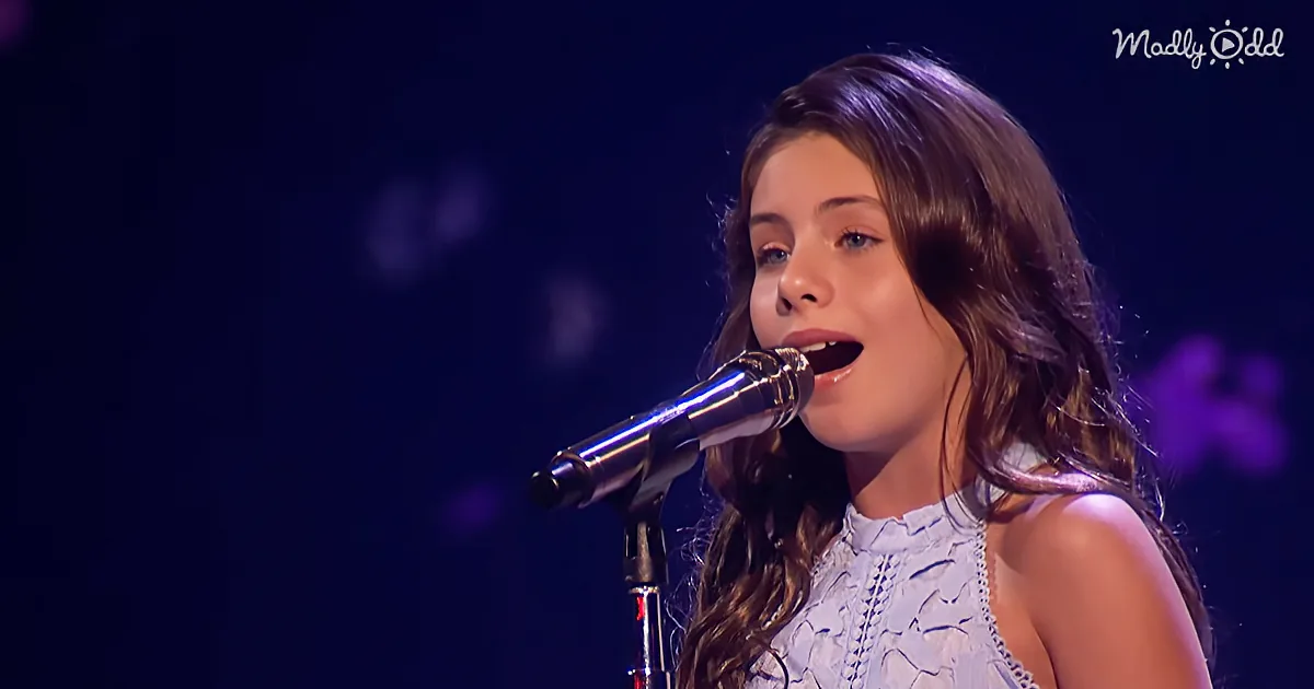 A young girl with long brown hair sings into a microphone on stage.