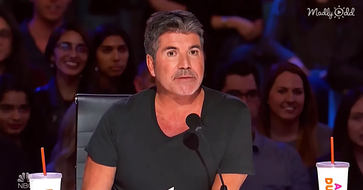 Simon Cowell sits at a judging panel with a microphone, surrounded by an audience.