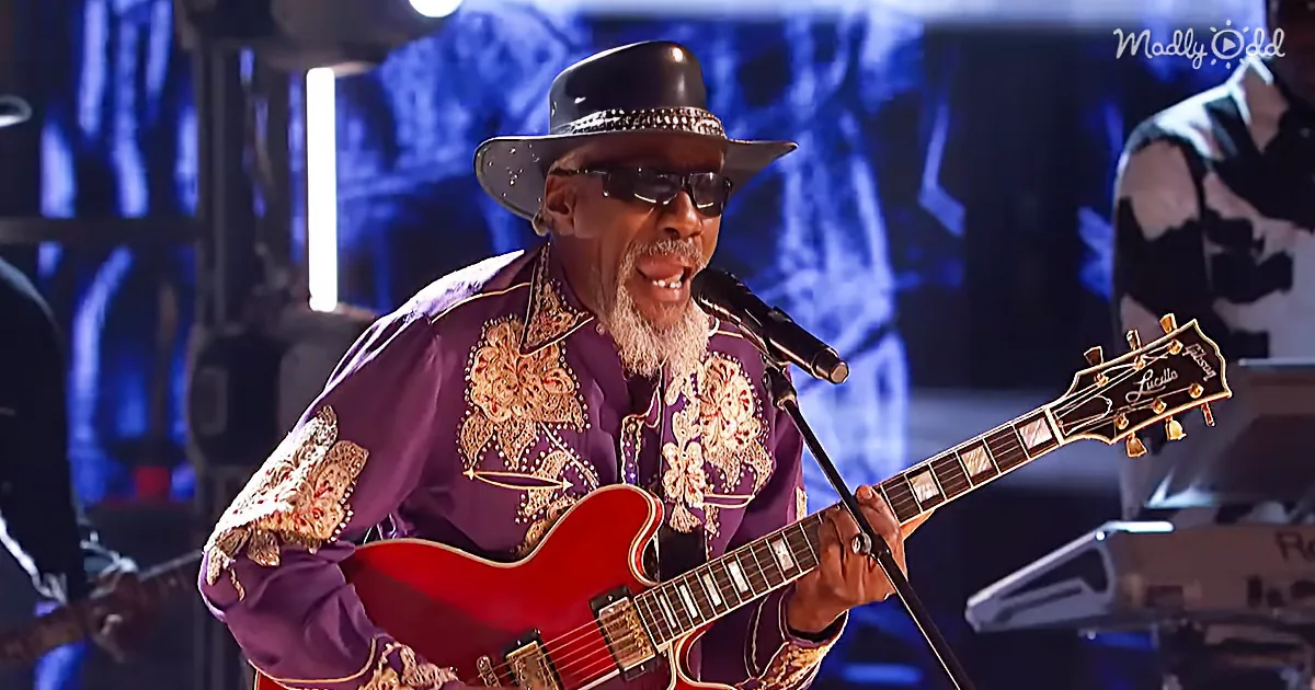 A man in a purple embroidered shirt and hat plays a red guitar on stage, singing into a microphone.