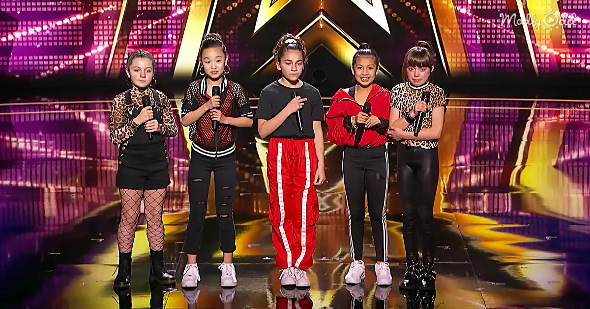 Five young performers stand on stage holding microphones, dressed in stylish outfits, with a vibrant background.