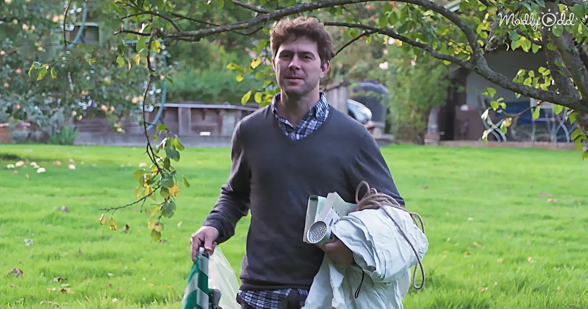 A man stands in a grassy yard holding a bag and a rolled-up item, with trees and a structure in the background.