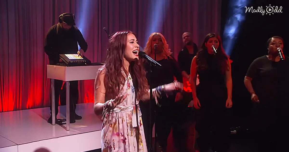 A woman in a floral dress sings passionately on stage with a band behind her.