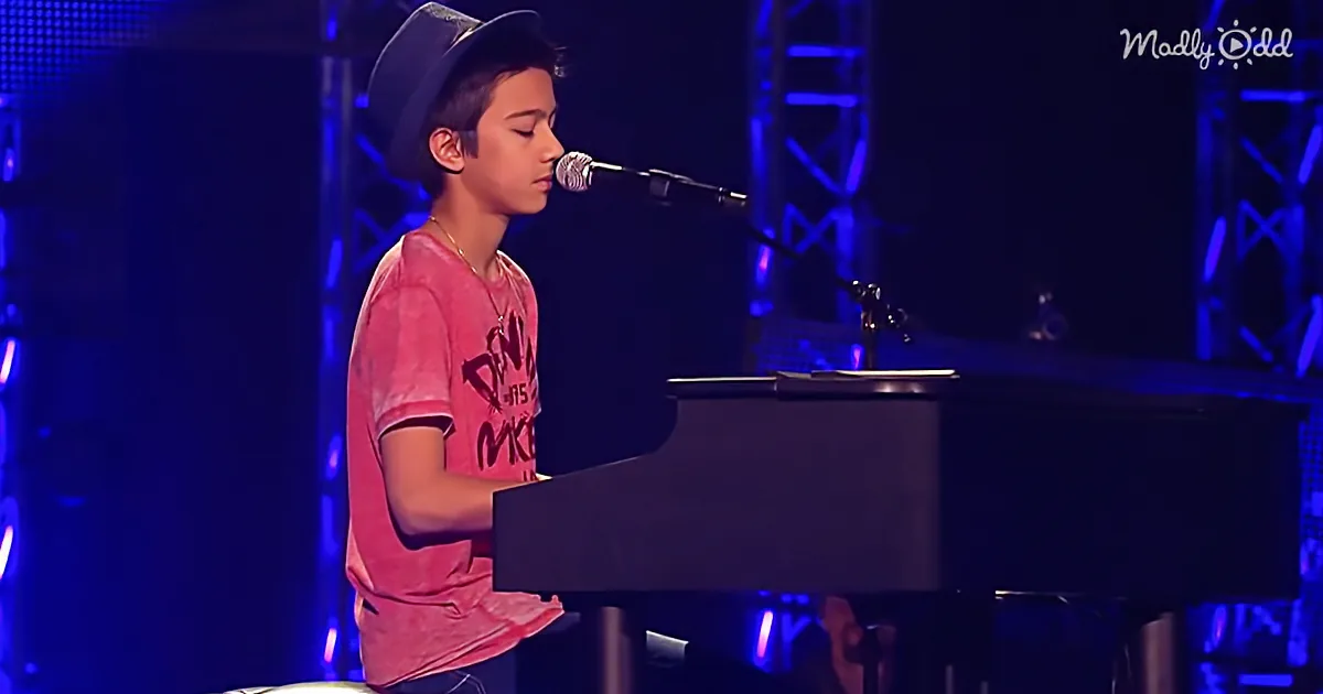 A young boy in a red shirt and hat plays piano on stage with a microphone.