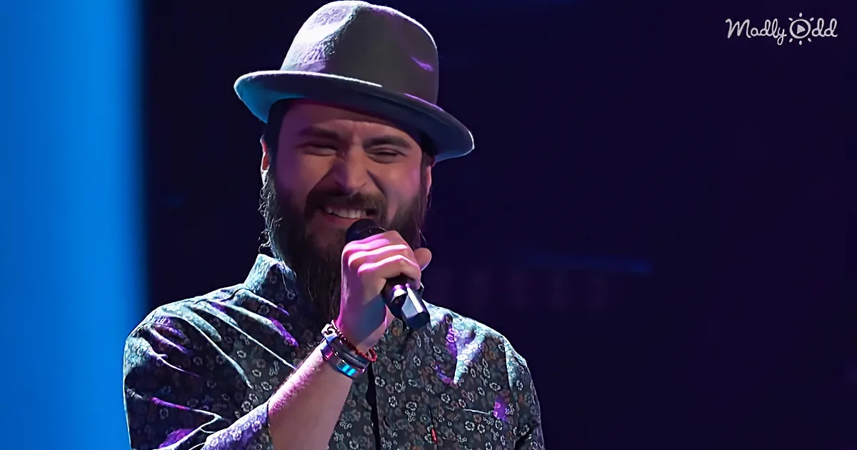 A man with a beard and hat smiles while holding a microphone on stage.