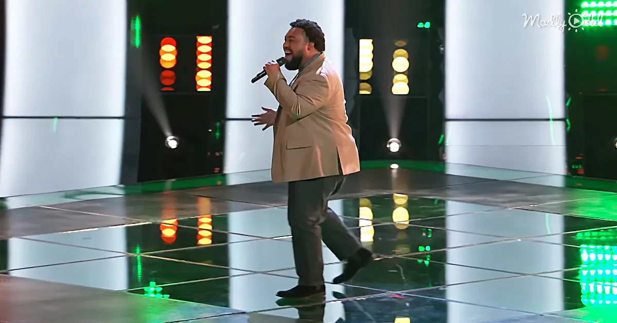 A man in a tan suit sings passionately on a stage with colorful lights and reflective floor.