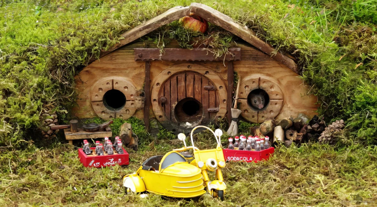 A miniature Hobbit house with a mouse, a yellow scooter, and soda crates in a grassy setting.