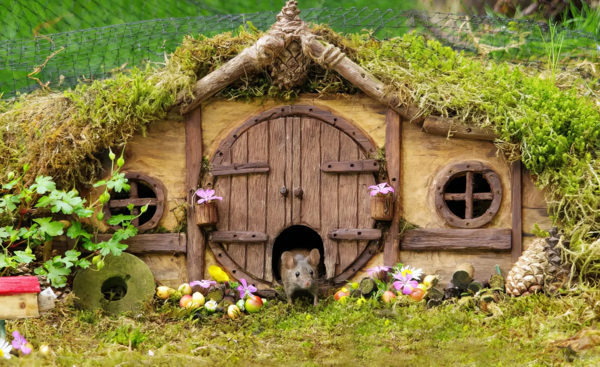 A small, moss-covered hobbit house with a round door and windows, surrounded by flowers and a mouse peeking out.
