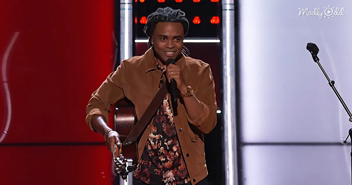 A man in a floral shirt and brown jacket holds a guitar and microphone on stage.