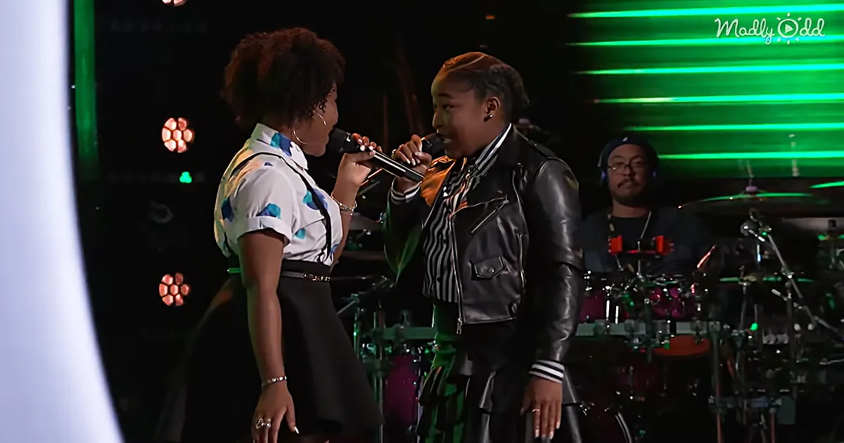 Two women sing on stage, one holding a microphone, with a drummer in the background.