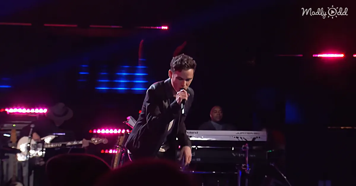 A man in a suit sings into a microphone on stage with colorful lights and musicians in the background.