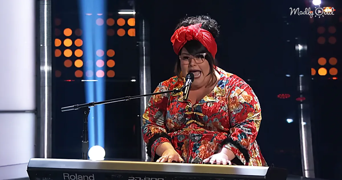 A person in a floral outfit and red headscarf sings into a microphone while playing a keyboard on stage.