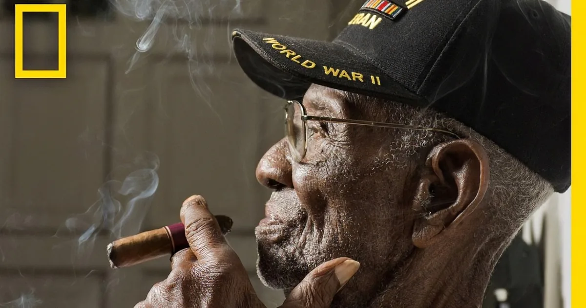 An elderly man wearing a "World War II" cap smokes a cigar, with smoke rising from it.