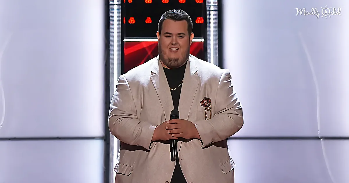 A man in a beige suit holds a microphone, smiling on a stage with red lights in the background.