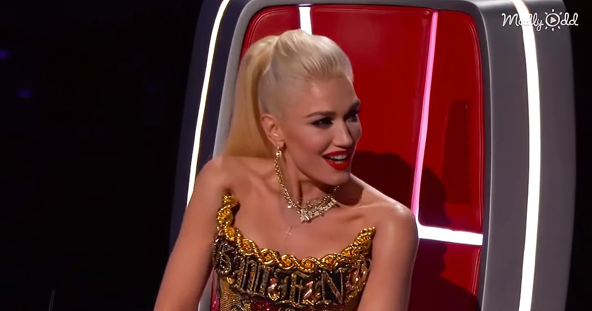 A woman with blonde hair in a ponytail, wearing a gold dress and necklace, sits in a red chair with white lights.