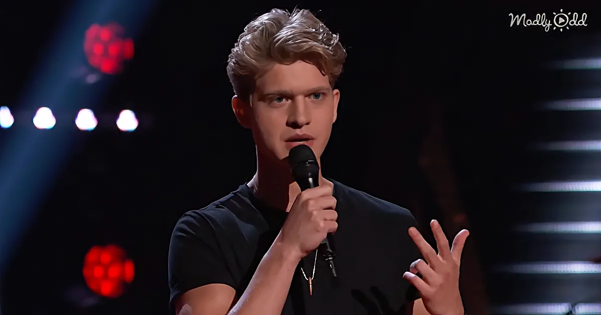 A young man with blonde hair, wearing a black shirt, holds a microphone and gestures with his right hand while speaking...
