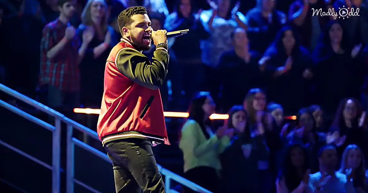 A man in a red and green jacket sings into a microphone on stage, surrounded by an audience.