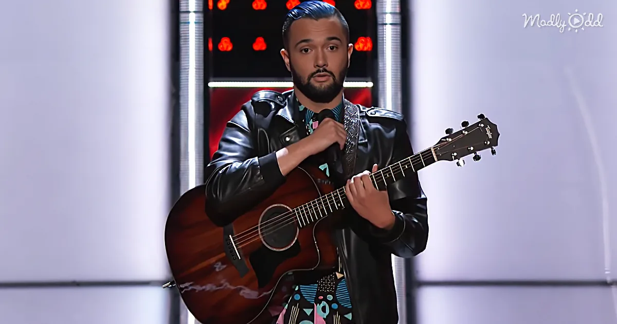 A man in a black leather jacket plays an acoustic guitar on stage with a microphone.