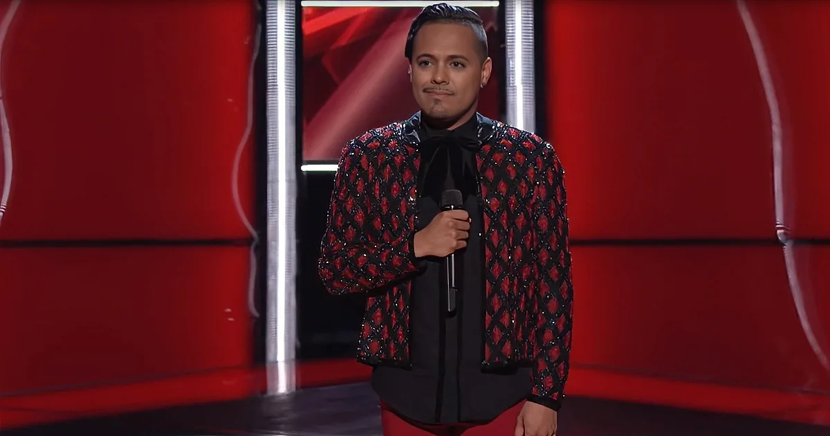 A man in a red and black patterned jacket holds a microphone on a stage with a red background.