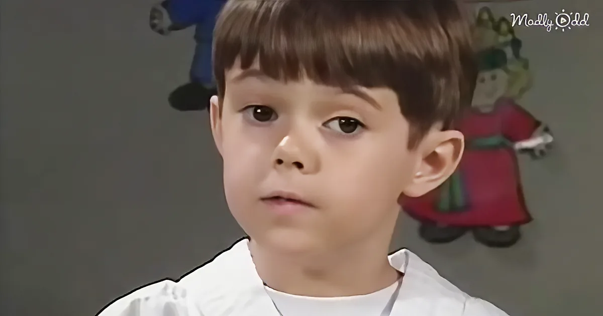 A young boy with short brown hair and bangs, wearing a white shirt, stands in front of a gray background with colorful...