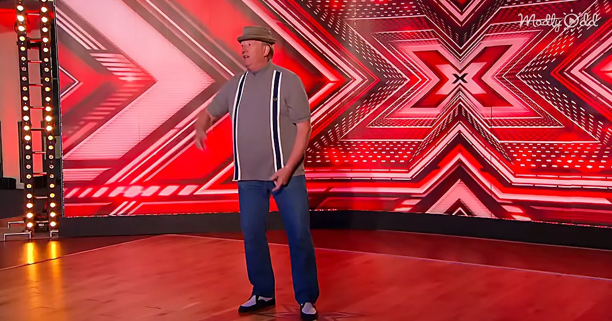 A man in a hat and suspenders stands on a stage with a red and black geometric background.