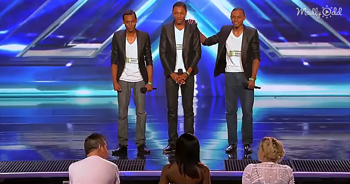 Three men in suits and white shirts stand on a stage with microphones, one has his arm around another.