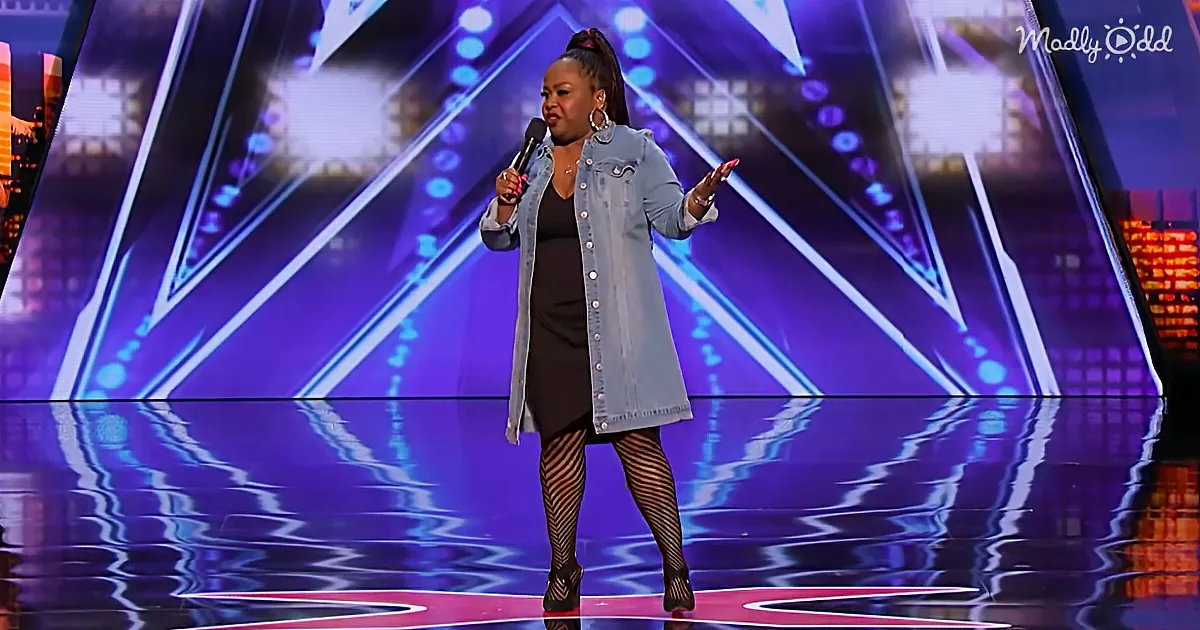A woman in a denim jacket and black dress stands on a stage, holding a microphone, with a vibrant purple background.