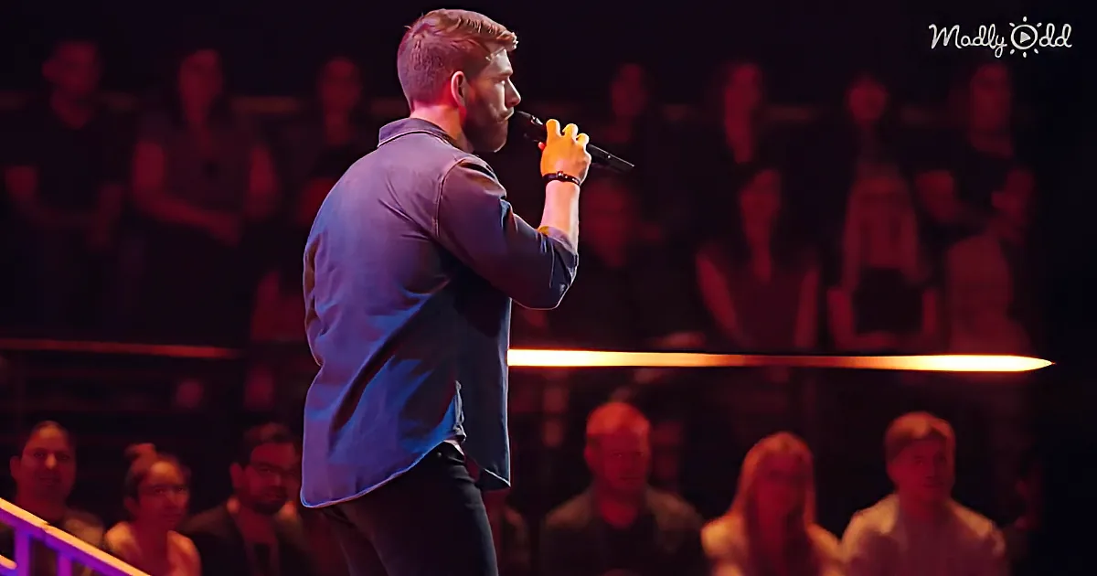 A man in a blue shirt holds a microphone on stage, with an audience in the background.