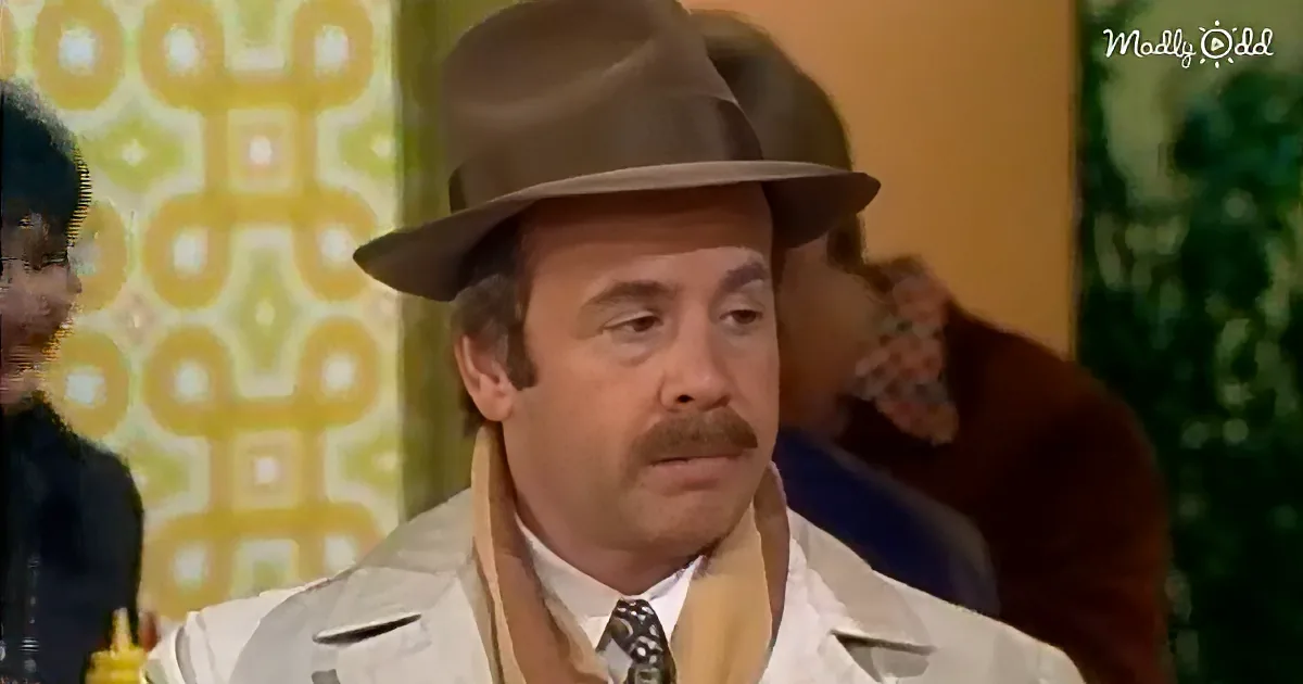 A man in a brown fedora and white shirt with a mustache looks to the side, with a patterned yellow wall and another...