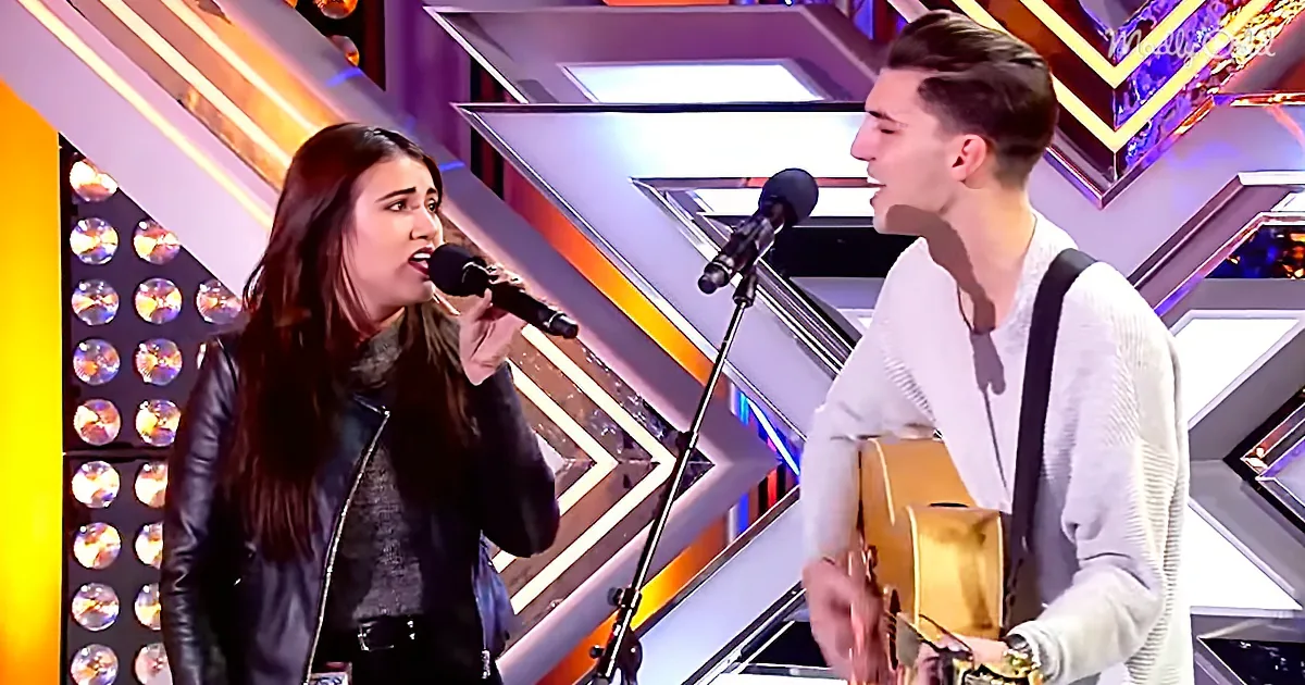 A woman sings into a microphone while a man plays guitar on a brightly lit stage.