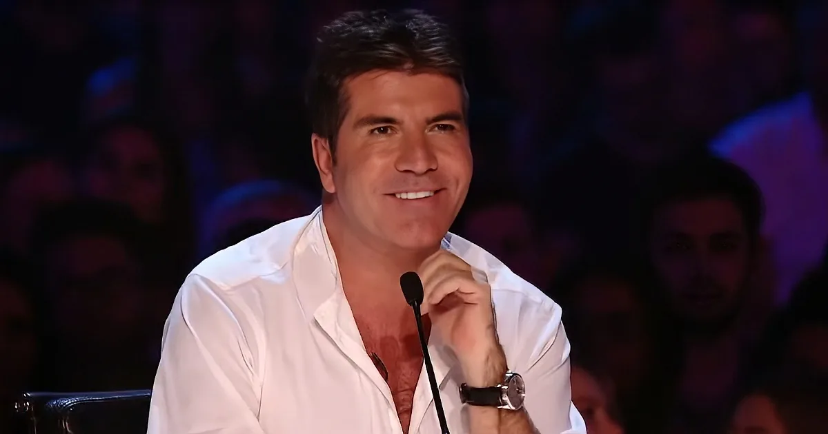 A man in a white shirt smiles, holding a microphone, with a blurred audience in the background.