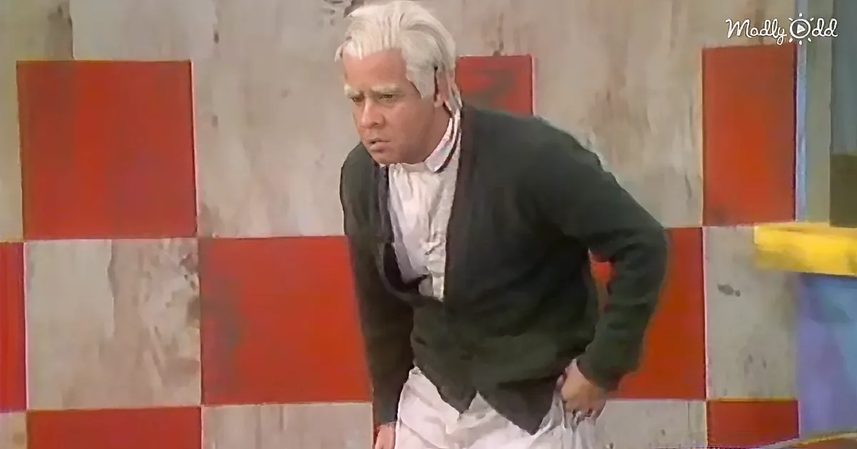 A man in a black jacket and white shirt leans forward, hands on knees, against a red and white checkered wall.