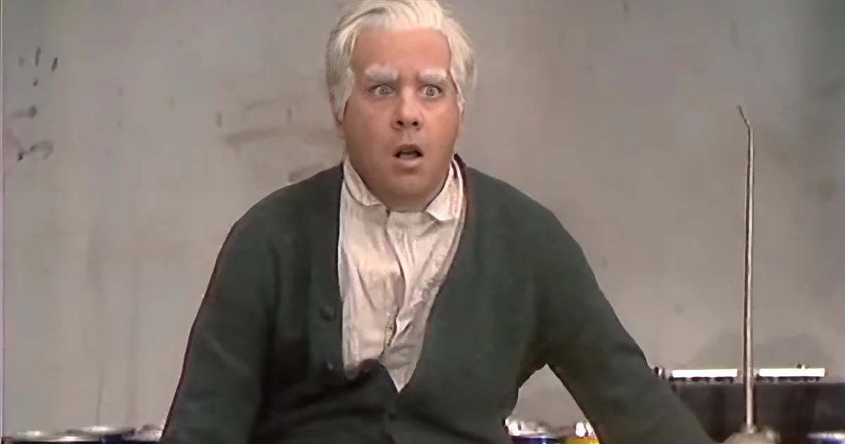 A man with white hair, wearing a green sweater and white shirt, appears surprised or shocked.