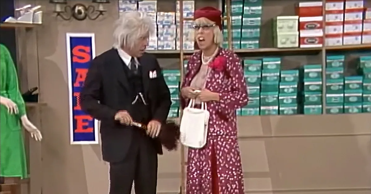 A man in a suit and a woman in a floral dress are standing in a store, seemingly engaged in a conversation.