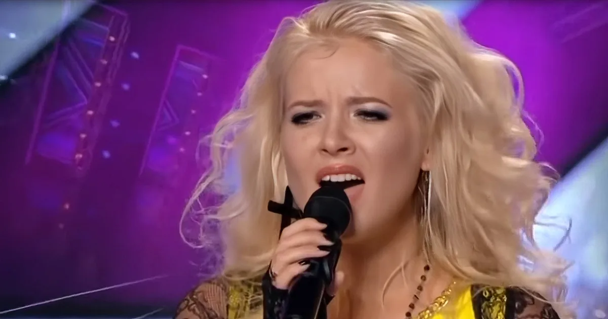 A woman with blonde hair sings passionately into a microphone on a stage with purple lighting.