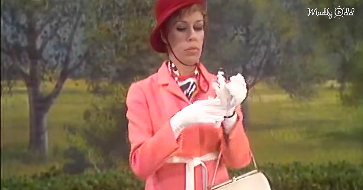 A woman in a pink outfit and red hat holds white gloves, standing outdoors with trees in the background.