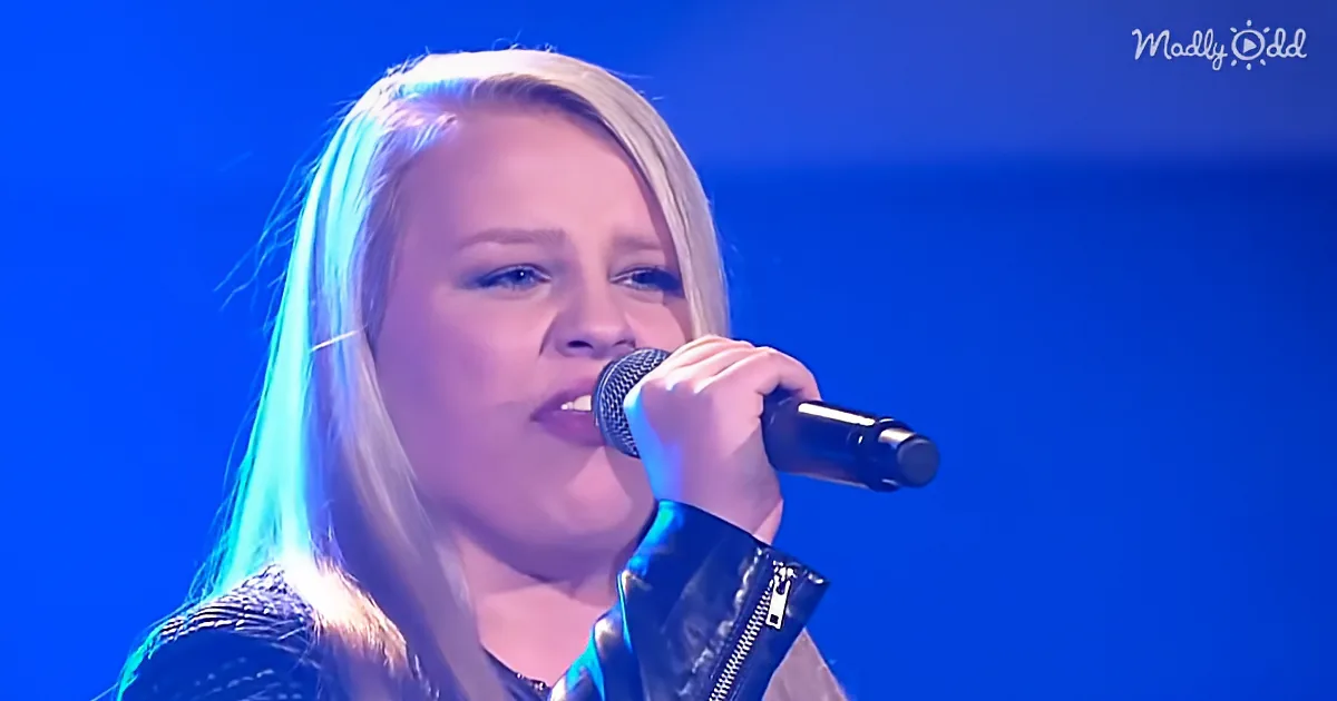 A person with long hair sings into a microphone against a blue background.