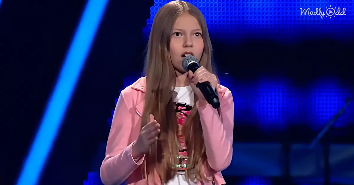 A young girl with long hair in a pink jacket sings into a microphone on a stage with blue lighting.