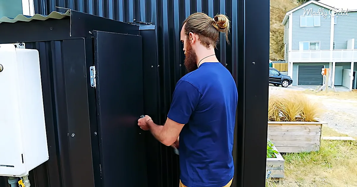 A man with a bun hairstyle and beard opens a black door, wearing a blue shirt.