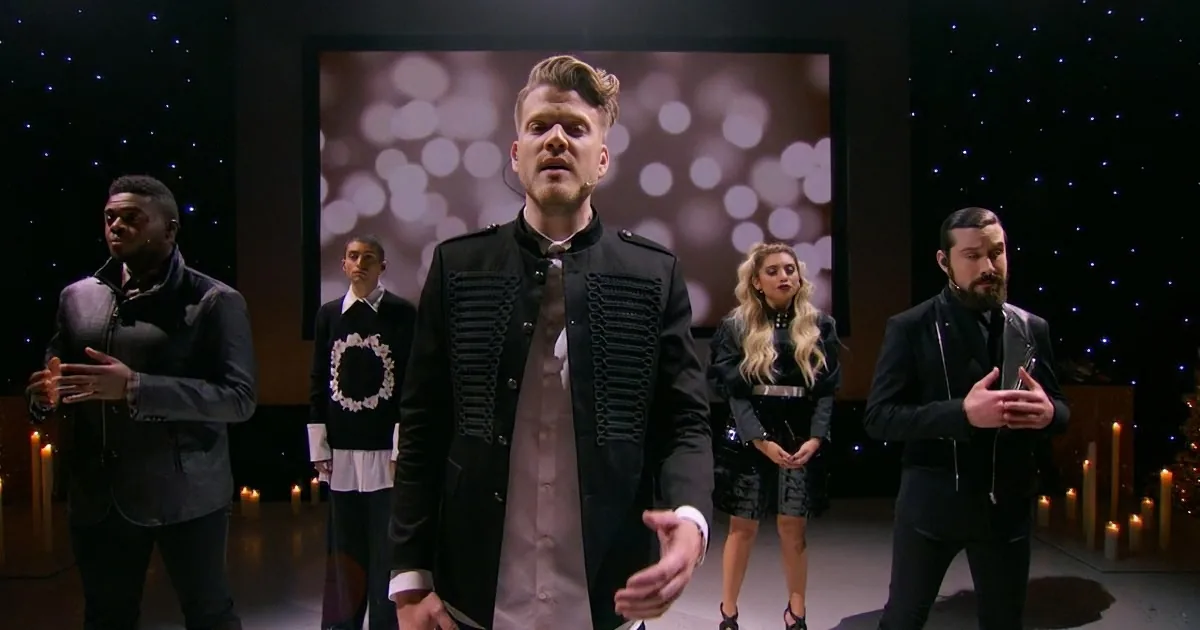 A man in a black jacket with buttons stands center stage, surrounded by others in formal attire, with a starry backdrop...