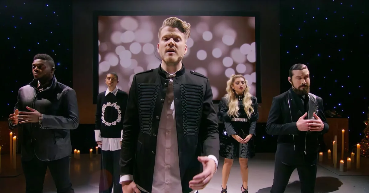 A man in a black jacket stands center stage, surrounded by others in similar attire, with a starry background and candles.