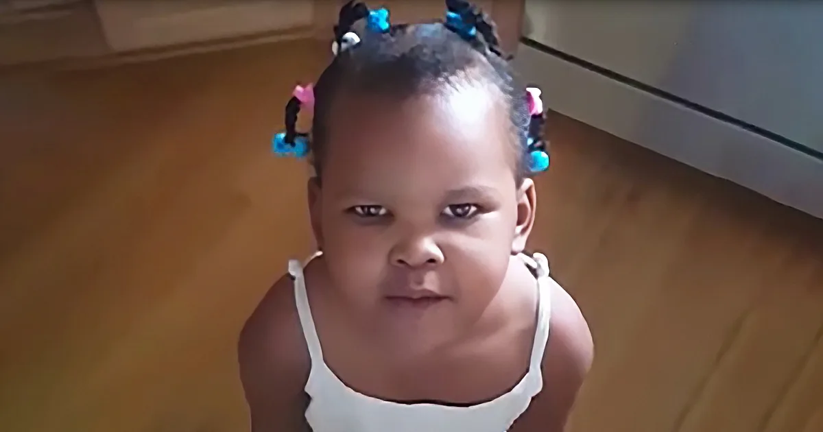 A young child with braided hair and a white top stands on a wooden floor.