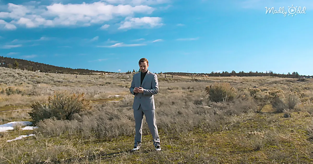 A man in a suit stands in a grassy, open field under a bright blue sky.